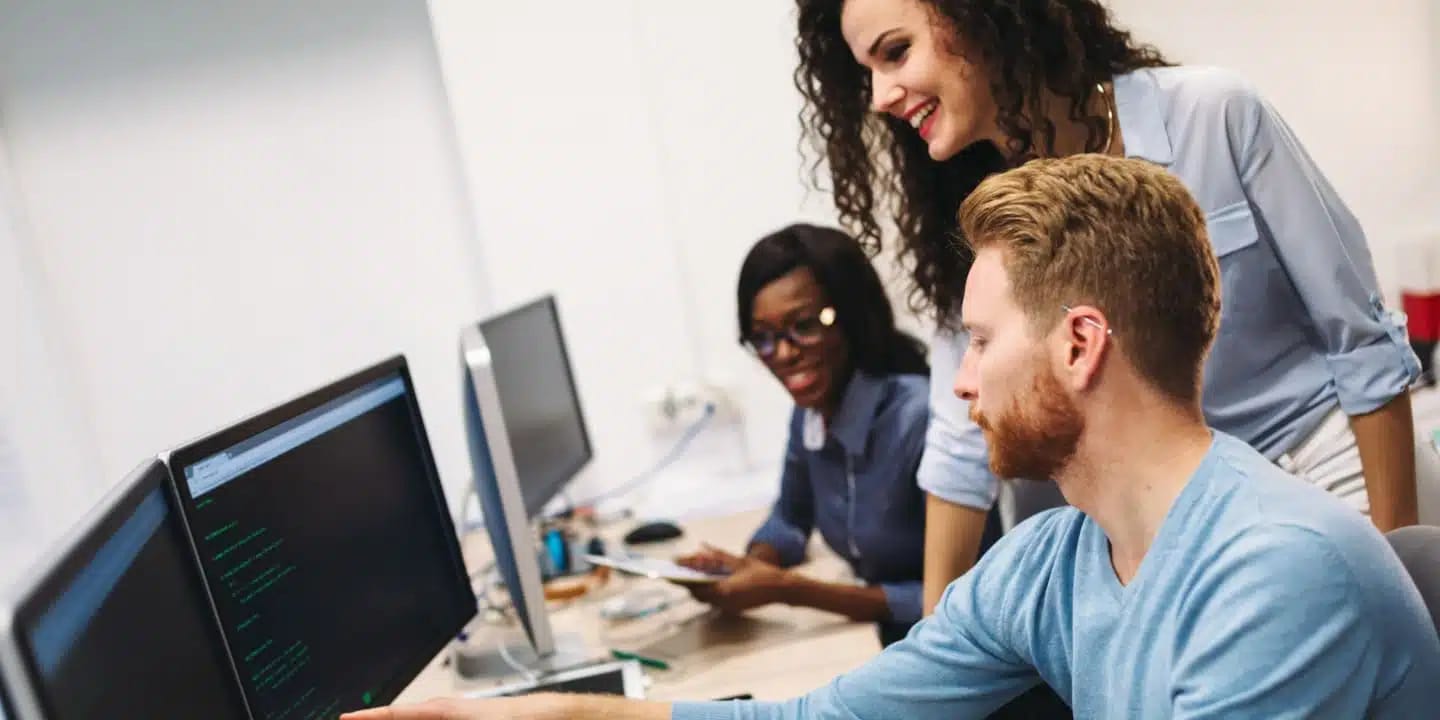 Three colleagues work together at computer desks; one man types while two women observe and discuss, all focused on code displayed on a monitor.