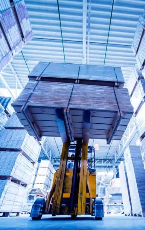 A yellow forklift lifts a large stack of lumber inside a warehouse with high ceilings and metal beams.