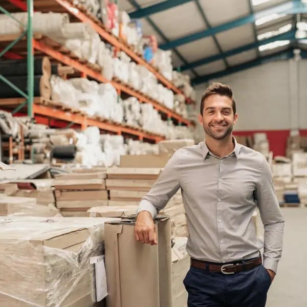 A man in a light gray shirt stands smiling in a warehouse filled with stacked boxes and shelves of materials.