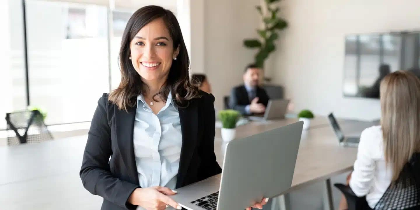 A woman in business attire holds a laptop and smiles in a modern conference room, with colleagues seated at a table in the background.