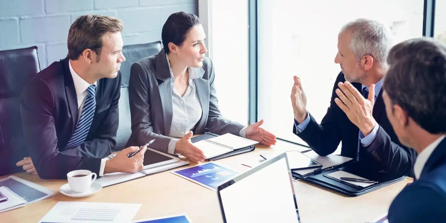 Four business professionals sit at a conference table engaged in discussion, with documents, notepads, and a laptop visible on the table.
