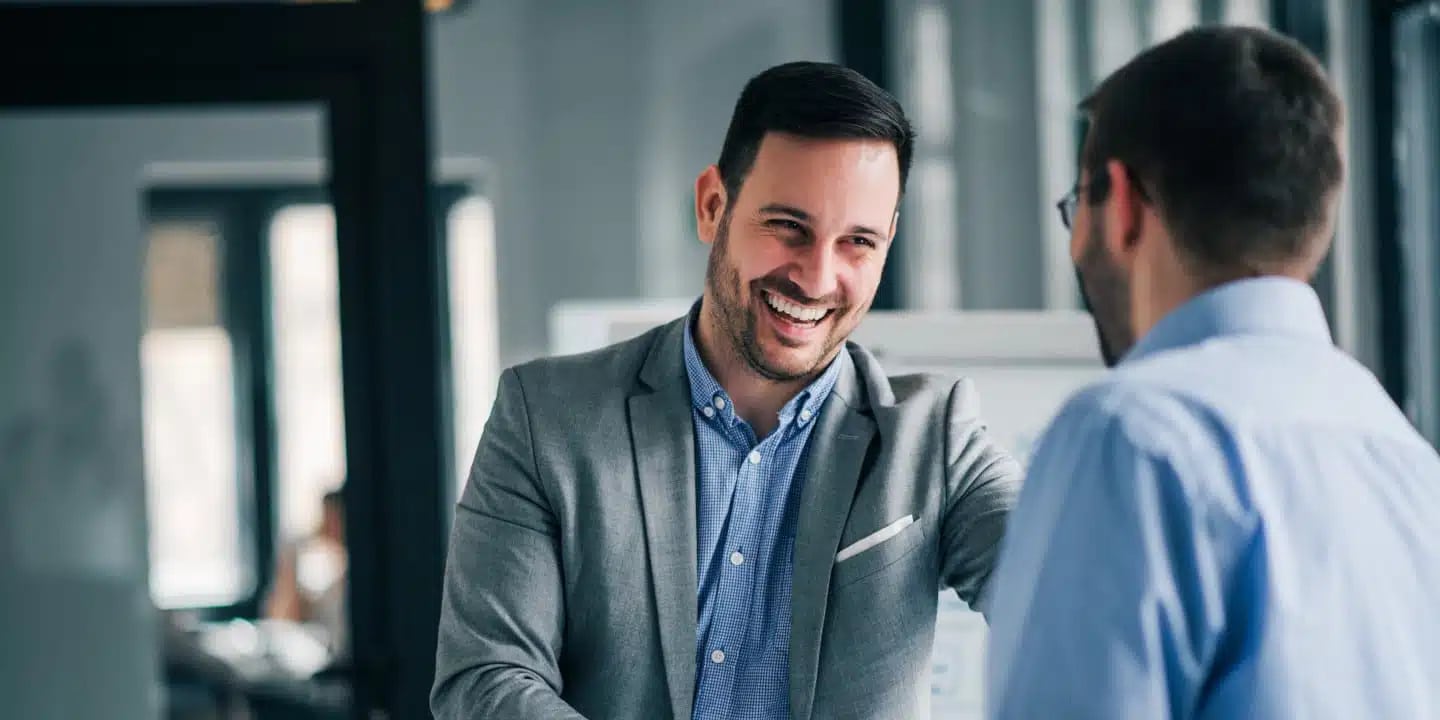 Two men in an office having a friendly conversation; one man is smiling and wearing a grey blazer, the other is seen from behind in a blue shirt.