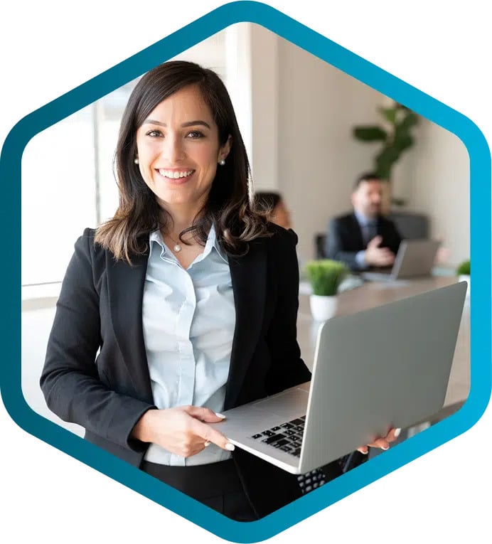 Woman in business attire holding a laptop, smiling, with colleagues working at desks in the background.