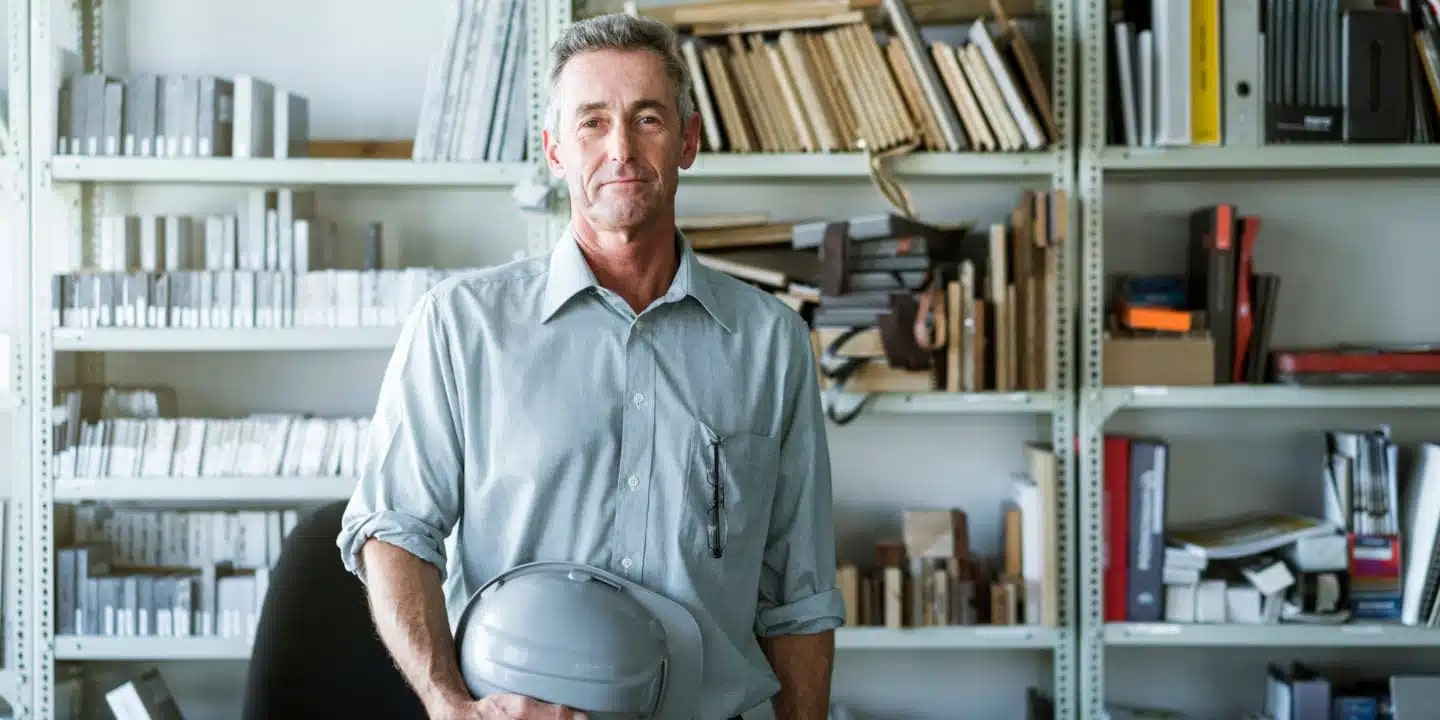 A man in a button-up shirt holds a hard hat and stands in front of shelves filled with books and files in an office or workspace.