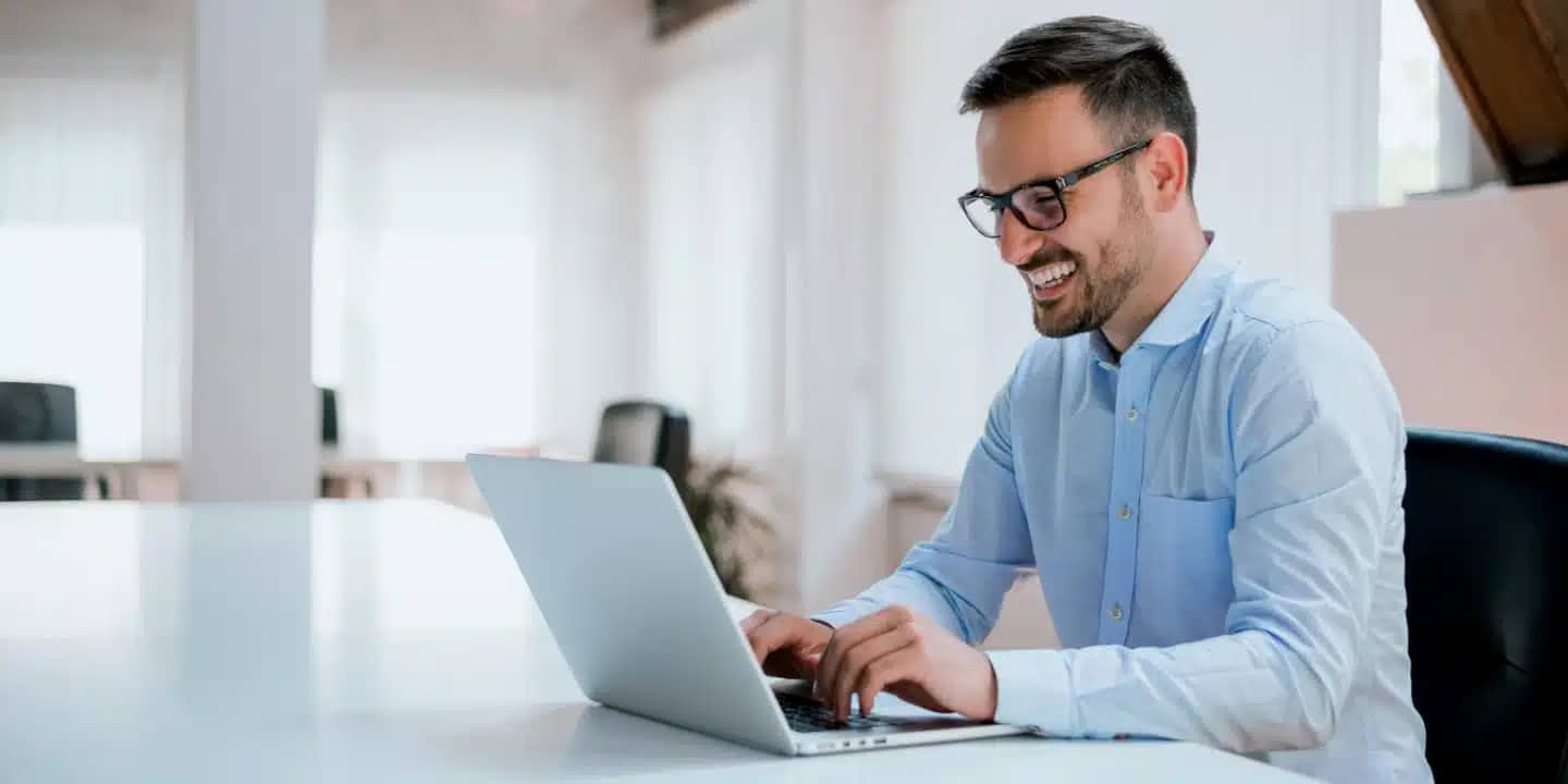 A man in a light blue shirt and glasses is smiling while typing on a laptop at a white desk in a bright office setting.
