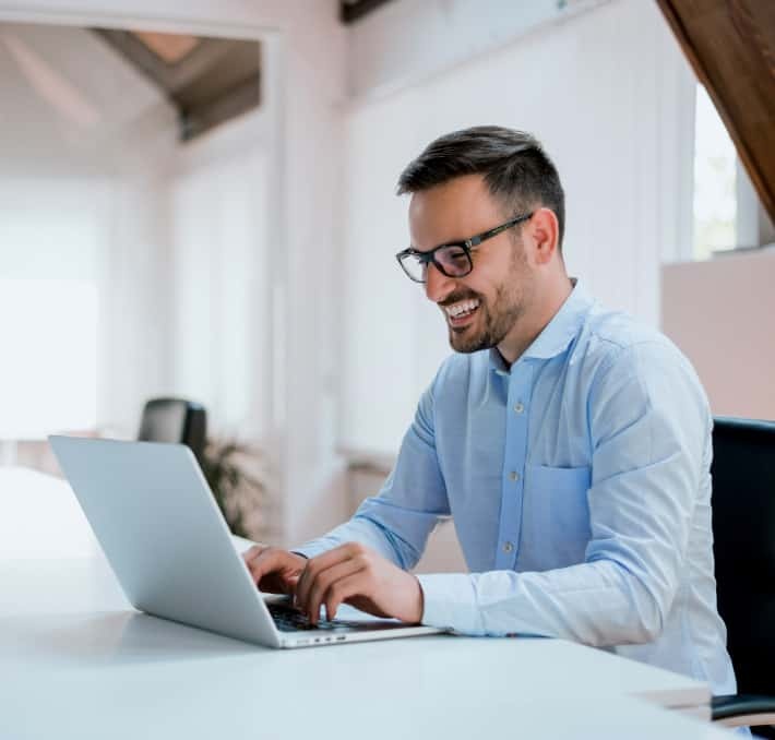 A man wearing glasses and a light blue shirt is smiling while using a laptop at a desk in a bright office space.