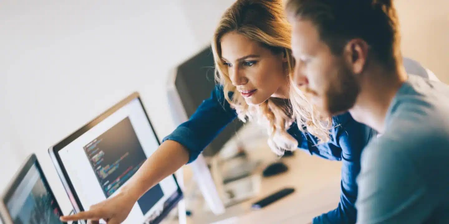 Two people working together at a desk, looking at computer screens with code displayed, as one person points at the monitor.