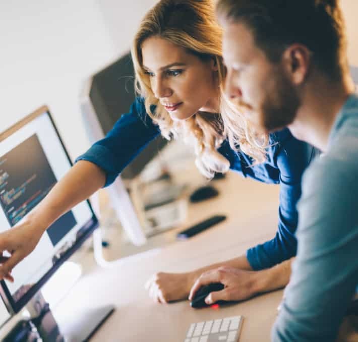 Two people work together at a computer, with one person pointing at code on the monitor while the other uses a mouse.