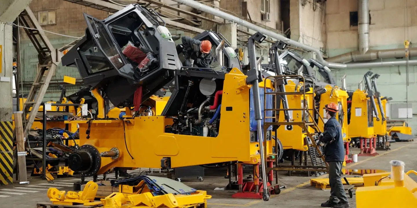 Workers in safety gear assemble and inspect large yellow industrial machinery inside a spacious factory setting.