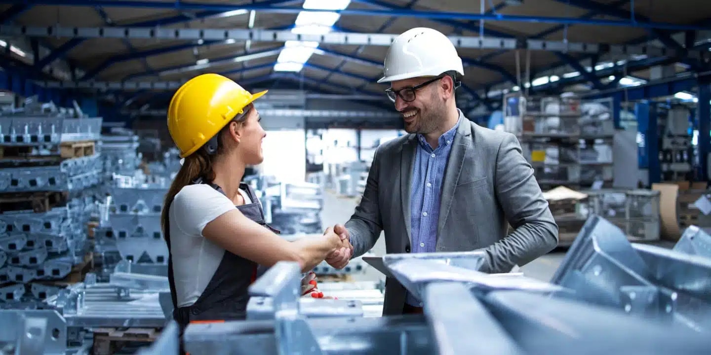 Two people in safety helmets shake hands and smile in a factory setting, surrounded by metal machinery and parts.