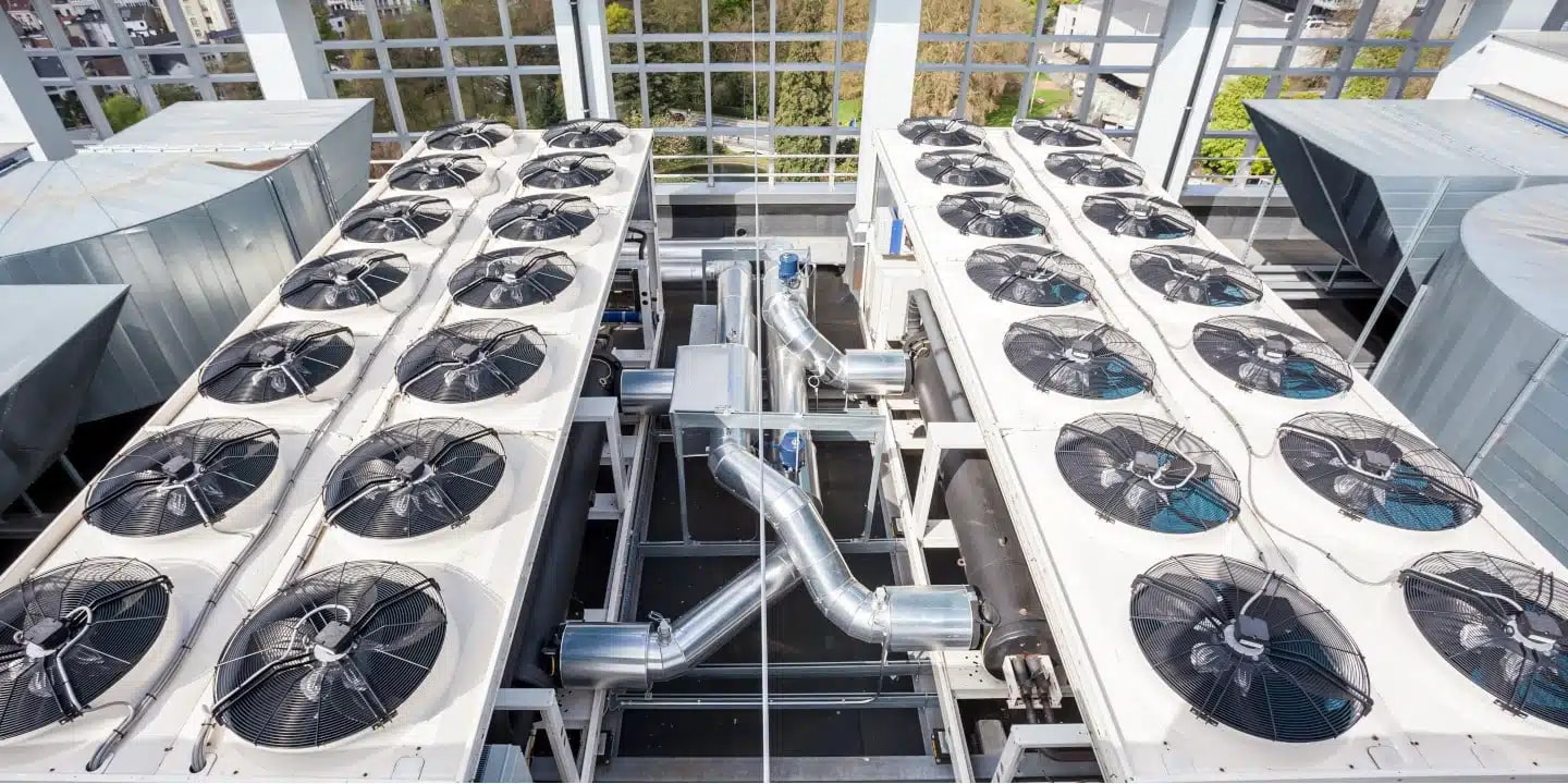Two large rooftop HVAC units with multiple cooling fans and metal ductwork, surrounded by a grid of windows with trees visible outside.