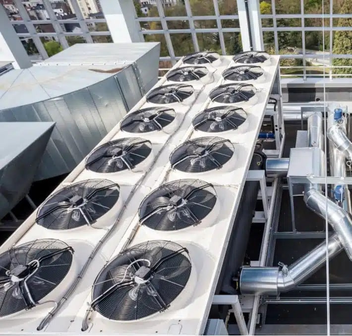 A rooftop HVAC system with multiple large industrial cooling fans and metal ducts, set against a glass building structure and a view of trees outside.
