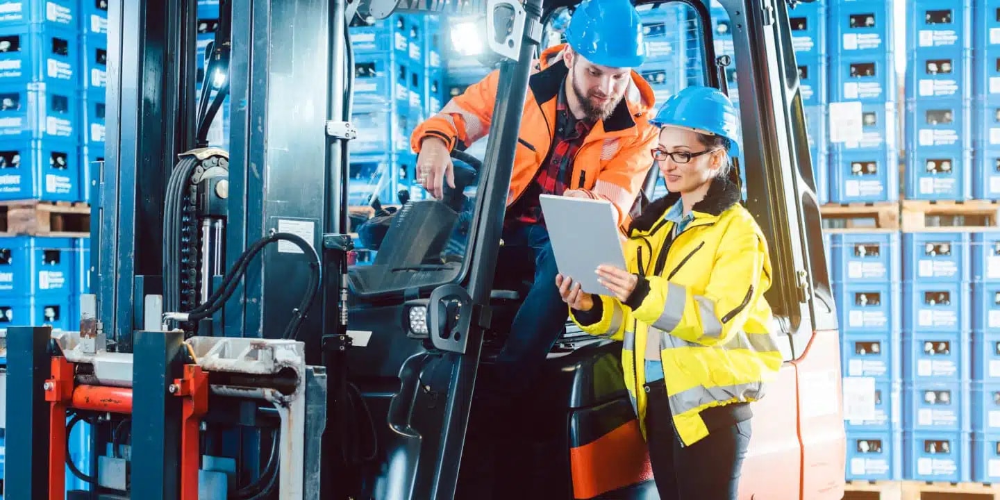 Two warehouse workers in safety gear review a clipboard next to a forklift, surrounded by stacks of blue storage bins in a warehouse setting.