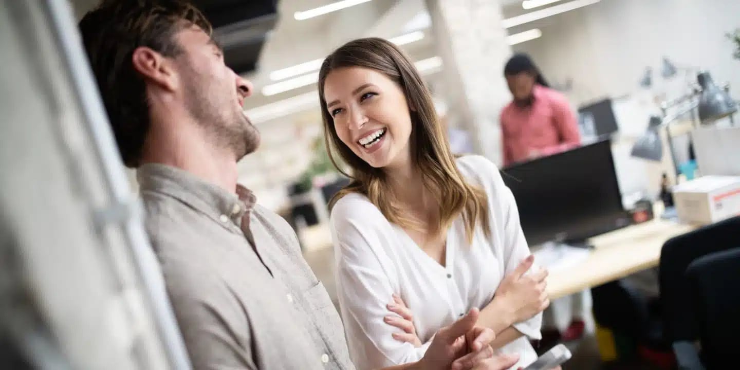 Two colleagues stand and chat, smiling and laughing together in a modern office space with desks and computers in the background.