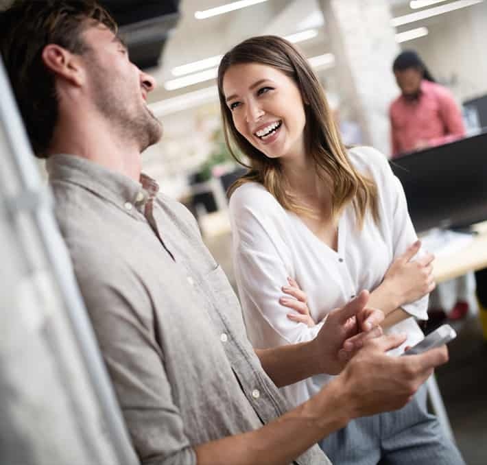 Two people stand and talk, smiling and laughing together in a modern office setting. One holds a smartphone. Other people work at desks in the background.