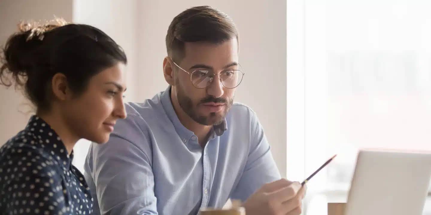 Two people sit at a desk looking at a laptop screen; the man points at the screen with a pen while the woman observes attentively.