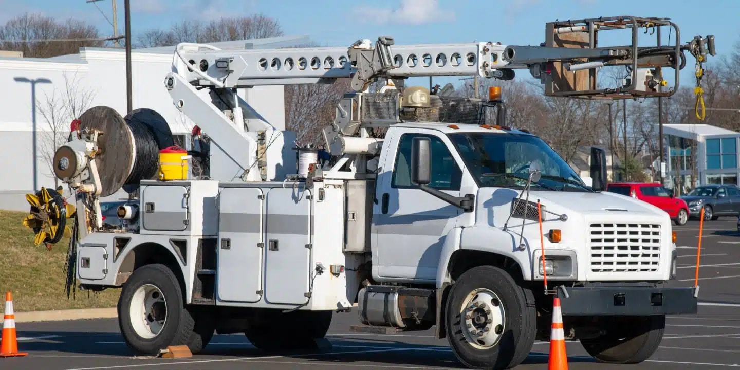 A white utility truck with a mounted boom lift is parked in a lot, with orange traffic cones around it and equipment stored on the trucks bed.