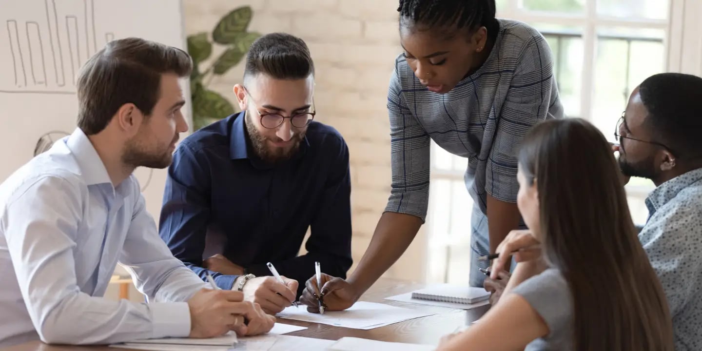 Five people sit around a table in an office, discussing documents and taking notes. One person stands and gestures towards the papers.