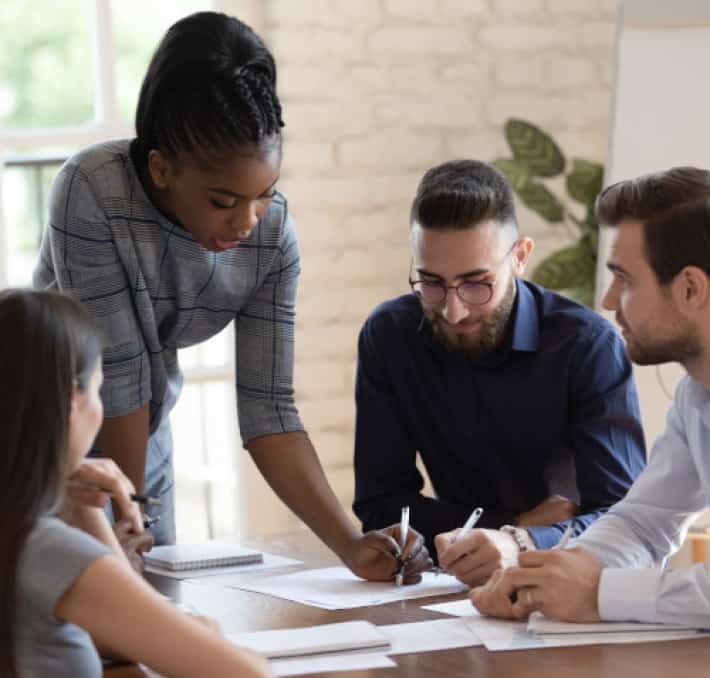 Four people sit around a table discussing documents; one woman stands and points at a paper while the others listen and take notes.