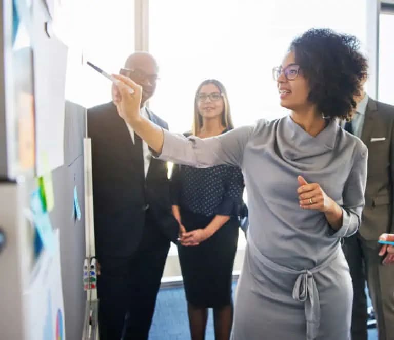 A woman points to a whiteboard while three colleagues stand nearby, observing her presentation in a brightly lit office setting.
