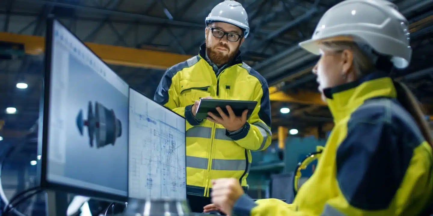 Two engineers in high-visibility jackets and hard hats review technical drawings on computer monitors in an industrial setting. One holds a tablet and takes notes.