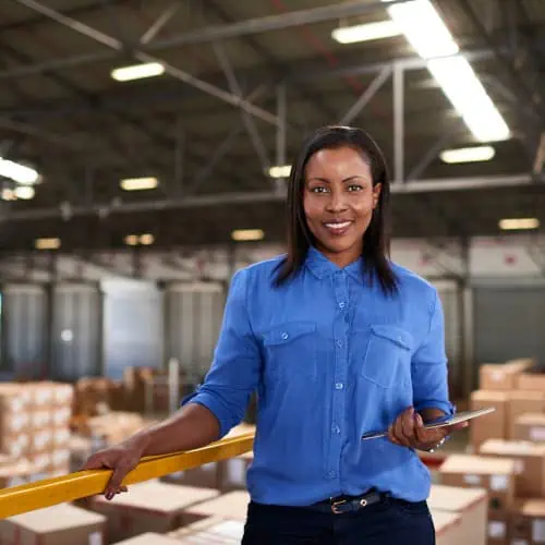 A woman in a blue shirt stands in a warehouse, holding a tablet and smiling, possibly using CPQ software to manage inventory, with shelves and boxes in the background.