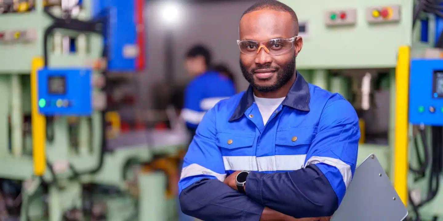 A man wearing safety glasses and a blue work uniform stands with folded arms in an industrial setting, holding a clipboard. Machinery and another worker are visible in the background.