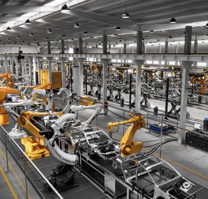 Automated robotic arms assemble car bodies on a production line inside a large, modern automotive factory.