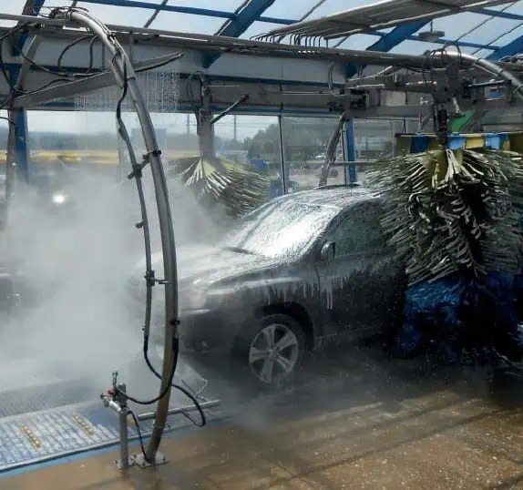 A black SUV is sprayed with water and soap as it passes through an automated car wash with rotating cleaning brushes.