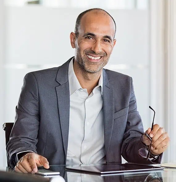A man in a gray suit sits at a desk, holding eyeglasses in one hand and smiling at the camera. A tablet is on the desk in front of him.