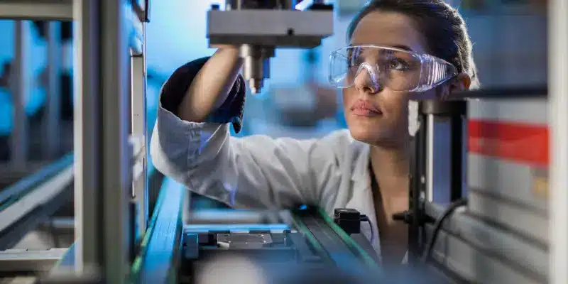 A woman wearing safety goggles and a lab coat operates or inspects equipment in a laboratory or industrial setting.