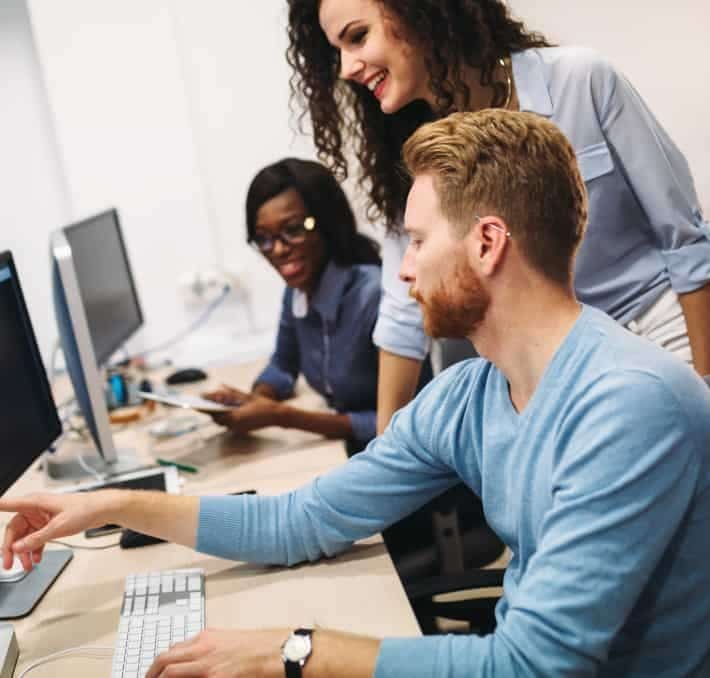 Three colleagues work together at computers in an office, with one man pointing at his screen while two women observe and discuss.