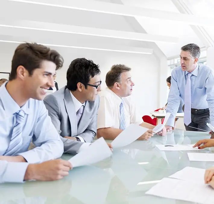 Four men in business attire sit at a conference table holding papers, while another man stands and speaks to them during a meeting in a bright office.