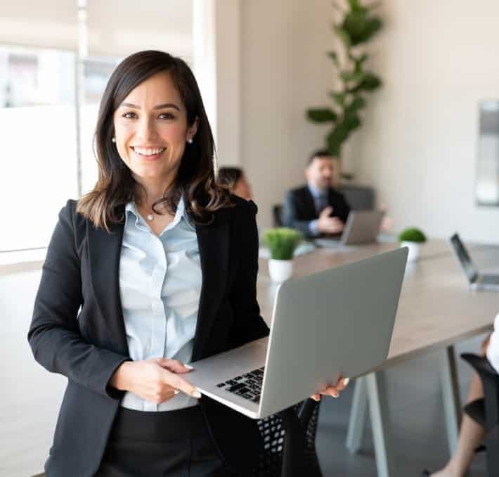 Woman in business attire holding a laptop and smiling, standing in a modern conference room with colleagues in the background.