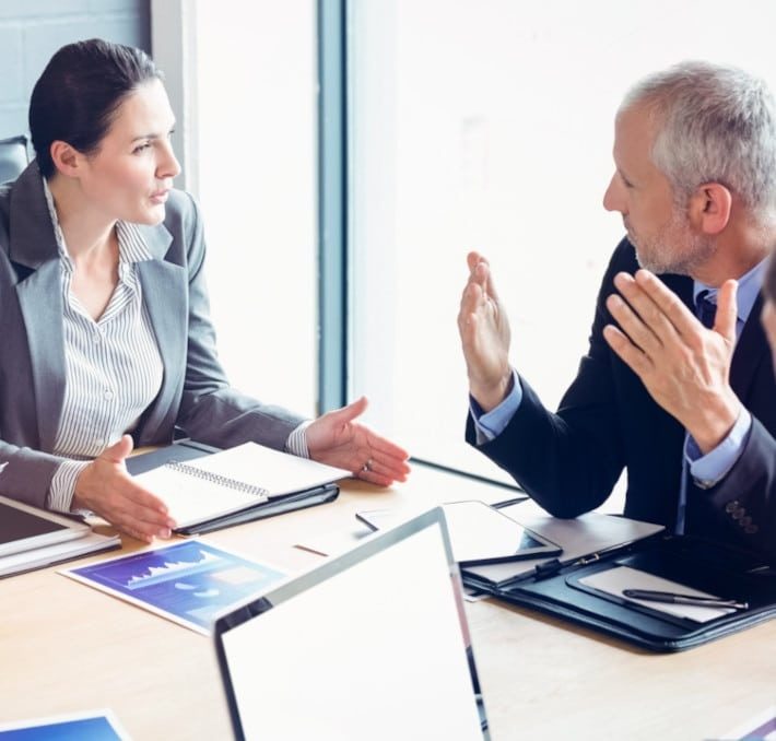 Two business professionals in formal attire have a discussion at a meeting table with documents, notebooks, and laptops in front of them.