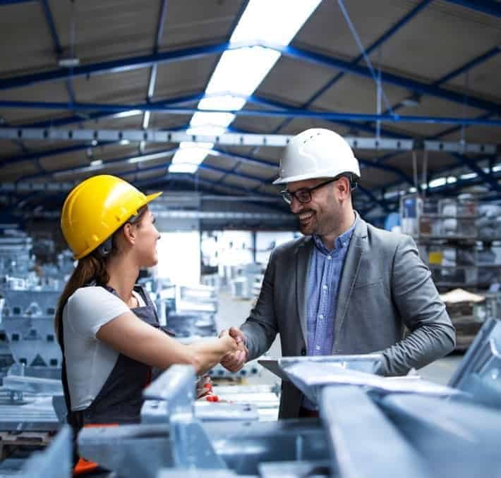 A woman in a yellow hard hat and overalls shakes hands with a man in a white hard hat and suit inside a factory or warehouse.