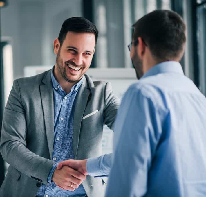 Two men in business attire shaking hands and smiling in an office setting.