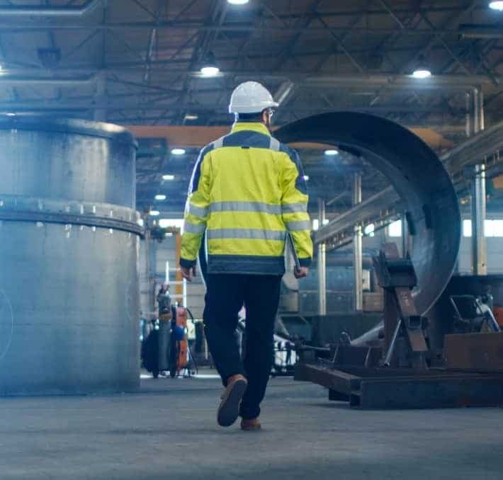 A person wearing a hard hat and high-visibility jacket walks through an industrial warehouse with large metal structures.