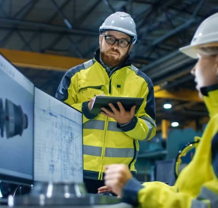 Two engineers in safety gear review technical drawings and 3D models on computer monitors in an industrial facility.