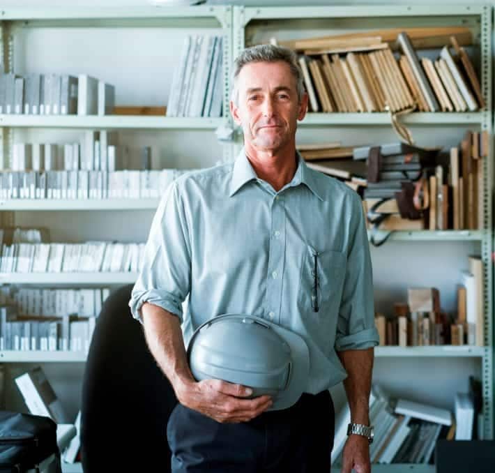 A middle-aged man in a collared shirt stands in front of shelves filled with books and files, holding a hard hat in one hand.