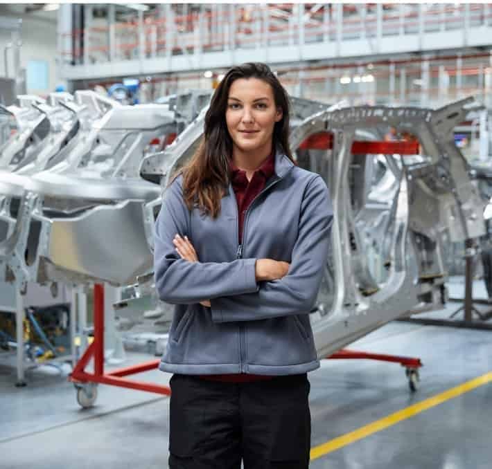 A woman stands with arms crossed in front of metal car frames in an automotive manufacturing facility.