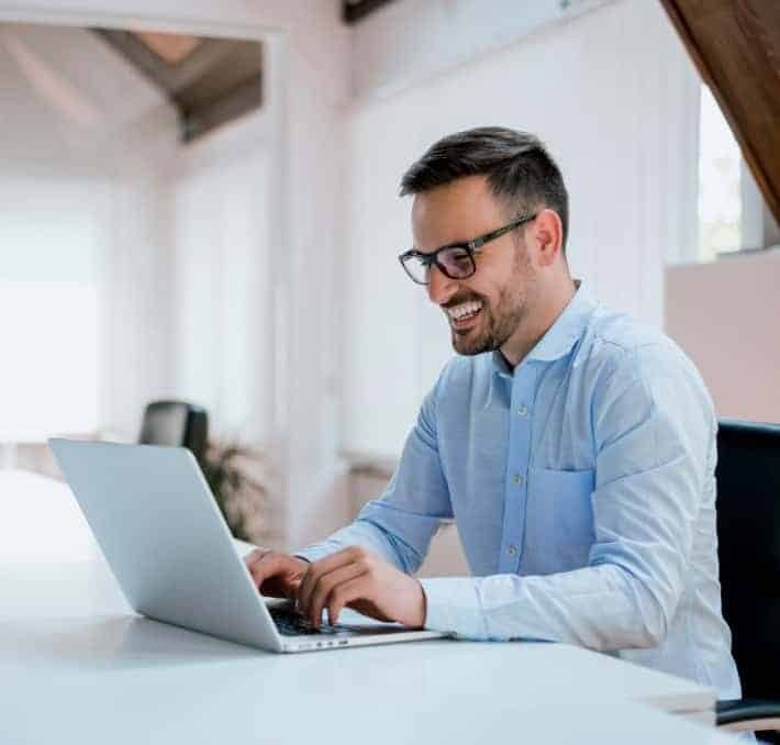 A man wearing glasses and a light blue shirt is smiling while using a laptop at a desk in a bright office space.