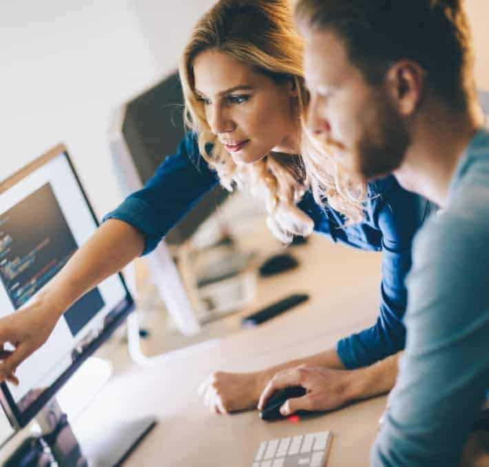 Two people work together at a computer, with one person pointing at code on the monitor while the other uses a mouse.