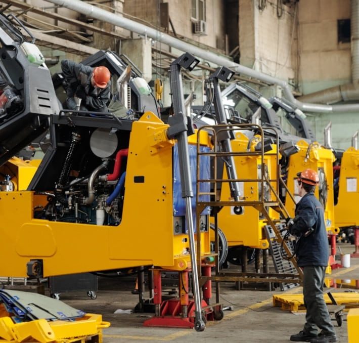 Two workers wearing safety gear assemble and inspect large yellow industrial machines inside a factory setting.