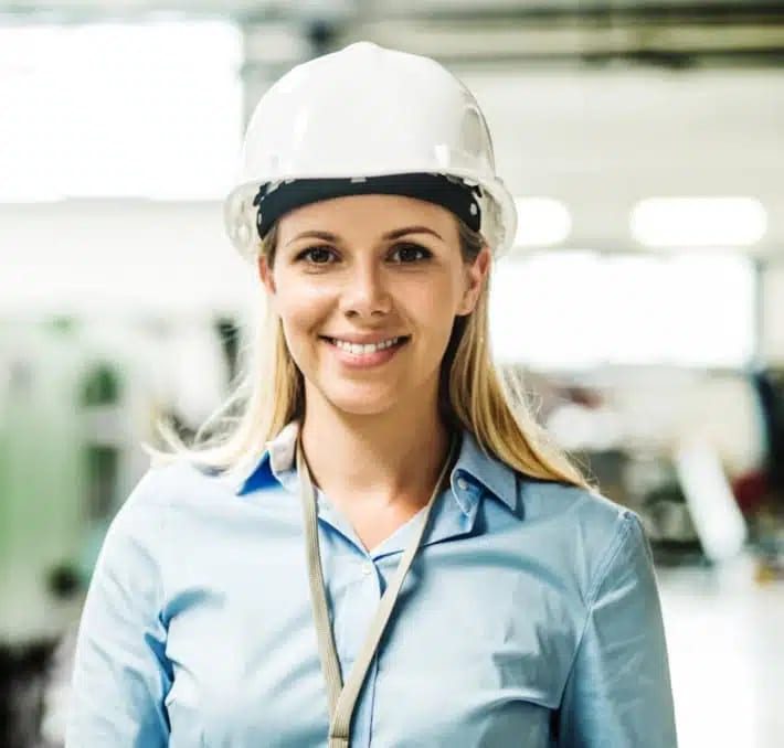 A woman wearing a white hard hat and a light blue shirt smiles at the camera in an industrial or workplace setting.