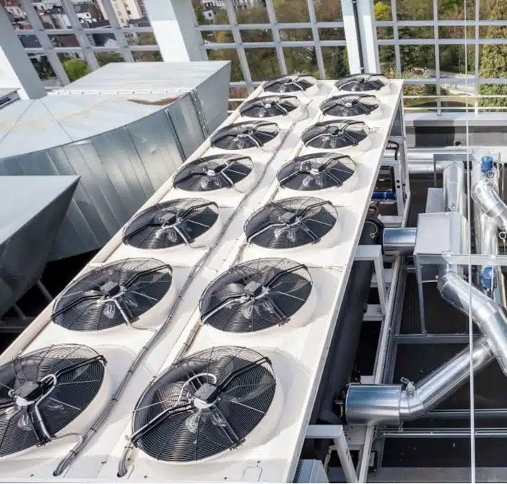 A rooftop HVAC system with multiple large industrial cooling fans and metal ducts, set against a glass building structure and a view of trees outside.