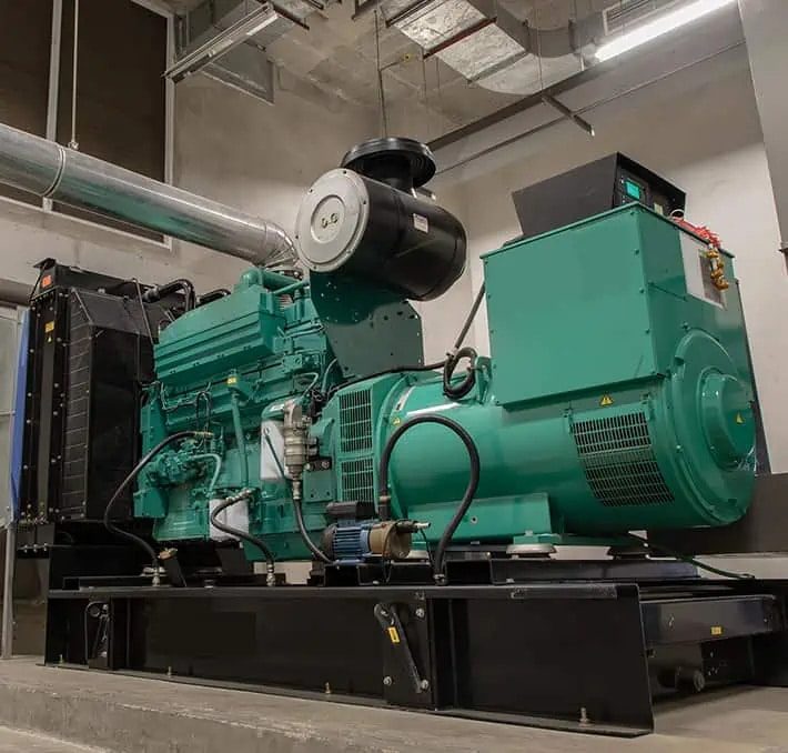 Large industrial diesel generator installed indoors on a concrete platform, surrounded by pipes and electrical wiring, with overhead lighting and visible ceiling infrastructure.