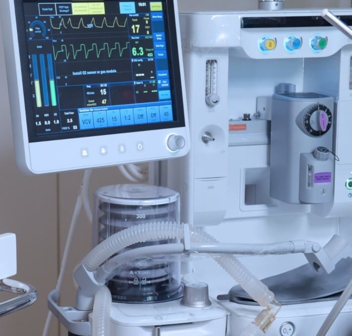 Close-up of a hospital anesthesia machine with a monitoring screen displaying patient vital signs and respiratory circuit tubing attached.