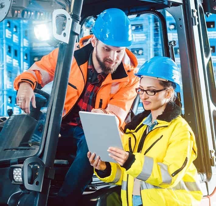 Two workers in safety gear and hard hats review information on a tablet next to industrial equipment in a warehouse setting.