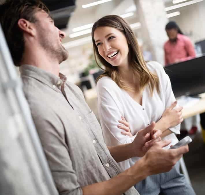 Two people stand and talk, smiling and laughing together in a modern office setting. One holds a smartphone. Other people work at desks in the background.